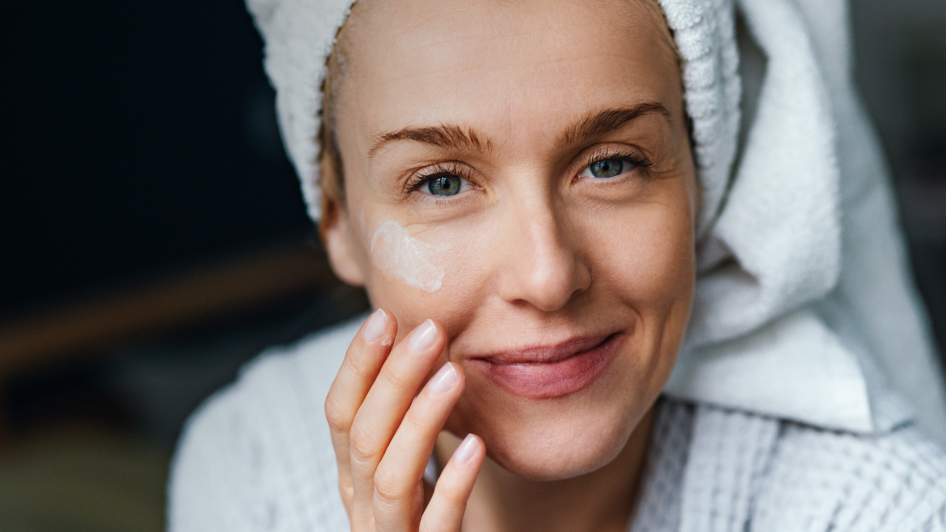 Eine entspannte Frau, mit einem Handtuch auf dem Kopf, trägt Gesichtscreme auf.